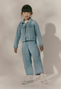 Lichtblauwe denimjas en bijpassende broek met verticale strepen. Voorzien van voorzakken en knopen als sluiting. Draagt combinatie met witte hoge sneakers.