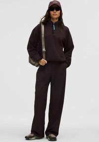 Donkerbruine fleece pullover met een halve rits, turquoise accent, gecombineerd met wijde donkere broek. Accessoires omvatten een donkere pet en zonnebril.