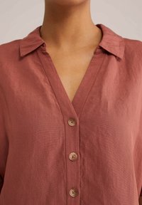 Rostfärgad linneskjorta med krage, med V-ringning och fem knappar framtill. Texturerad tyg med synliga sömnadsdetaljer.
