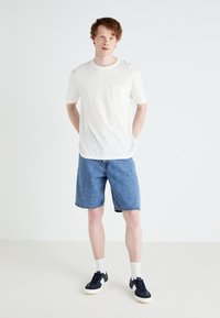 Fehér, rövid ujjú póló zsebével, kék farmer rövidnadrággal párosítva. A modellen sötét sportcipő van fehér talppal és bokazoknival.