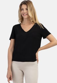 Tricou negru cu mânecă scurtă și decolteu în V, confecționat dintr-un material moale și neted, având o croială lejeră și un tiv ușor curbat, purtat cu pantaloni deschiși la culoare.