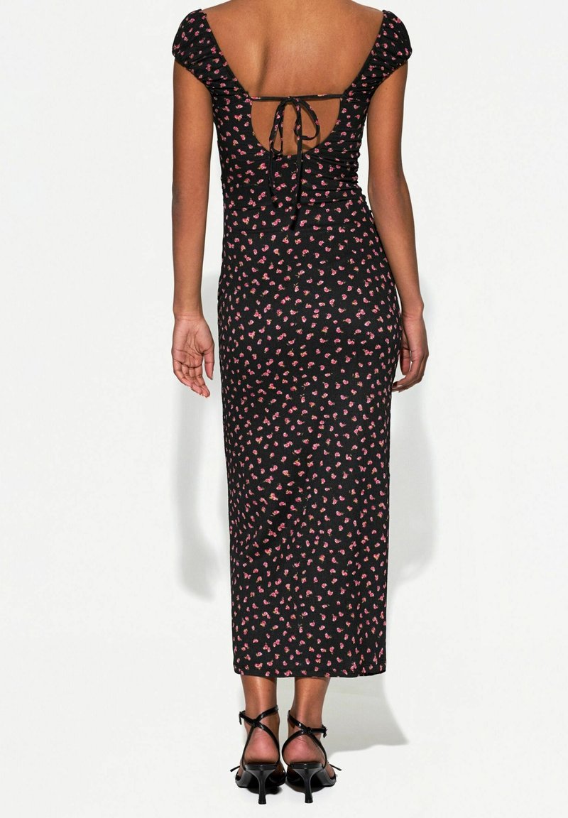 Femme portant une robe midi noire à petites fleurs roses, dos nu noué avec des cordons, et des sandales à talons noires avec des lanières.
