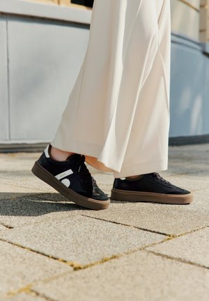 Schwarze Sneakers aus Wildleder und Leder, mit einer braunen Gummisohle und weißen Akzenten, getragen zu crèmefarbenen weit geschnittenen Hosen.