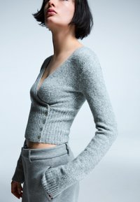 Szürke kötött kardigán mély V-nyakkal, bordázott textúrával és oldalsó gombos részletekkel. Hosszú ujjak enyhén bővülő mandzsettával.