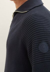 Maglione zip-up blu navy con spalle a costine testurizzate e corpo liscio. Presenta una patch rotonda con logo sulla spalla sinistra.