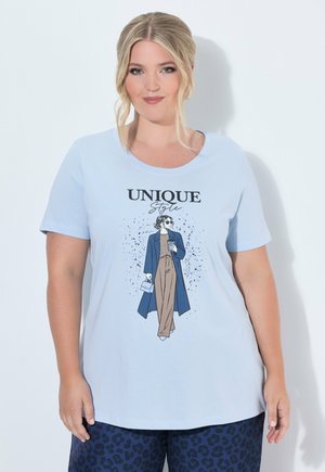 Donna con capelli biondi che indossa una maglietta azzurra con il testo "Unique Style" e un'illustrazione di una donna alla moda che tiene una borsa e un caffè.