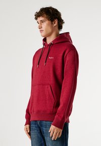 Bordo spalvos mikropluošto džemperis su kišenėmis, reguliuojama kapuce, bedugnio rankovėmis ir maža emblema.