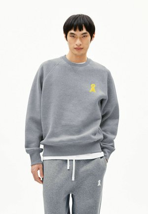 Junger Mann trägt ein graues Sweatshirt und eine Jogginghose mit kleinen gelben und weißen Logos, steht vor einem schlichten weißen Hintergrund.