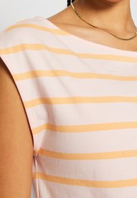 Gros plan d'une personne portant un haut rose clair avec des rayures horizontales orange et un collier fin en or.