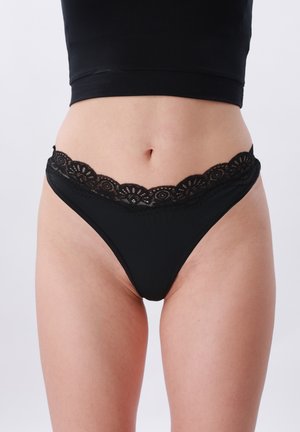 Kobieta nosząca czarne majtki z koronkową lamówką o wysokim kroju oraz czarny krótki top, pokazana od talii do górnej części ud na jednolitym tle.