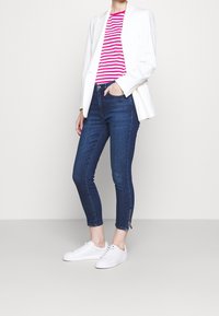 Jeans ajustados de color azul oscuro con cintura de altura media, cremalleras en los tobillos, combinados con una blazer blanca y una camisa a rayas rosa. Zapatillas blancas completan el look.