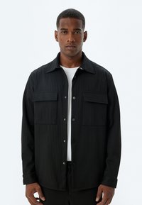 Chaqueta de camisa a rayas negras con dos bolsillos en el pecho, hecha de un tejido texturizado. Presenta un cierre frontal con botones y un diseño de cuello clásico.
