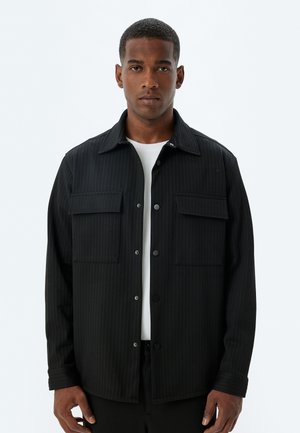 Giacca camicia a righe nere con due tasche sul petto, realizzata in un tessuto strutturato. Presenta una chiusura con bottoni e un design classico del colletto.