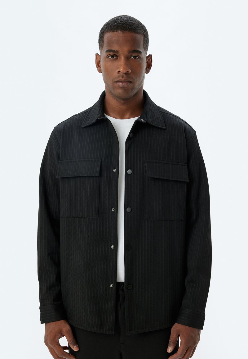 Chaqueta de camisa a rayas negras con dos bolsillos en el pecho, hecha de un tejido texturizado. Presenta un cierre frontal con botones y un diseño de cuello clásico.