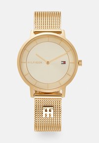 Goldfarbene Uhr mit rundem Gehäuse und minimalistischem elfenbeinfarbenem Ziffernblatt. Verfügt über ein Maschenarmband und ein dezent graviertes Logo auf der Schnalle.