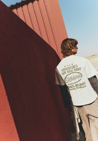 Hvid t-shirt med korte ærmer, der har grøn tekstprint med teksten "Eventyr Begynder Her I Dag Oklahoma" og en lagdelt langærmet skjorte under.