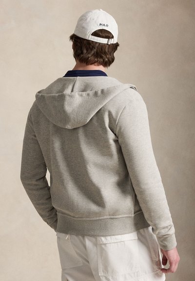 Homme de dos portant un sweat à capuche gris clair, une casquette Polo blanche, un col de chemise bleu marine et un pantalon cargo blanc sur un fond beige uni.