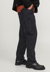Zwarte cargobroek met meerdere zakken, een ontspannen pasvorm en een duurzaam katoenmateriaal. Gepaard met zwarte schoenen en een oranje shirt eronder.