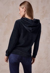 Felpa con cappuccio a coste scure, dal tessuto morbido, abbinata a jeans blu scuro. Il design presenta una vestibilità ampia e maniche lunghe.