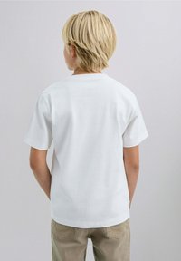 Blond kind met de rug naar de camera, gekleed in een eenvoudig wit T-shirt met korte mouwen en beige broek, tegen een neutrale achtergrond.