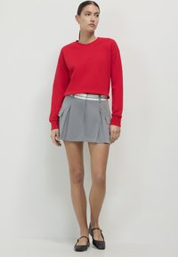 Felpa rossa a maniche lunghe abbinata a una gonna plissettata grigia con tasche laterali. Il modello indossa delle ballerine nere con dettaglio a cintura.