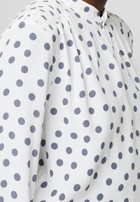 White blouse with navy blue polka dots, featuring a stand collar and gathered detailing at the shoulders for added texture.