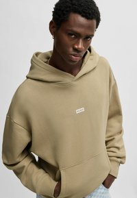 Beige hoodie in een gestructureerde stof. Bevat een wit label met "HUGO." Boxy ontwerp met een voorzak en een ontspannen pasvorm.