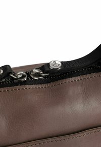 Bruine lederen tas met een zwarte rits, zilverkleurige hardware en een ankeraccentknopje aan de ritstrekkker. Textuur oppervlak en nette stiksels.