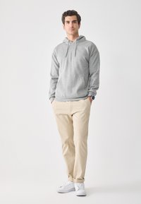 Joven de pie con las manos en los bolsillos, vistiendo una sudadera gris, pantalones beige, zapatillas blancas y un reloj de pulsera, frente a un fondo blanco liso.
