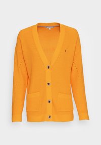 Keltainen neulecardigan, jossa on V-pääntie, kaksi etutaskua ja suuret ruskeat napit. Siinä on röyhelöinen kuvio ja rento istuvuus.