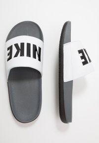 Nike šlepetės su balta sintetine viršutine dalimi, dideliu juodu logotipu, pilku padu ir tekstūruotu juodu padu, užtikrinančiu sukibimą.