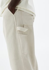 Pantalons en coton beige avec une finition texturée, dotés de poches latérales et arrière, et d'un accent de sangle détaillé sur la poche avant.