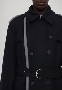 Cappotto doppiopetto blu navy con dettagli grigi, colletto a risvolto e fibbia circolare argentata. Presenta grandi bottoni e spalline.