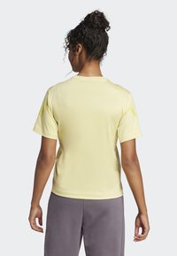 Žluté tričko s krátkým rukávem a kulatým výstřihem, hladká textura a kontrastní žluté detaily na rukávech. Nosené s šedými kalhotami.