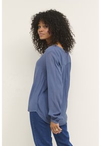 Långärmad blå topp i en texturerad tyg, med rund halsringning och avslappnad passform, ihop med blå denimbyxor.