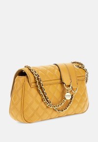 Sennepsgul quilted læderhåndtaske med gylden kædetras og cirkulært Guess-logo charme på frontflappen.