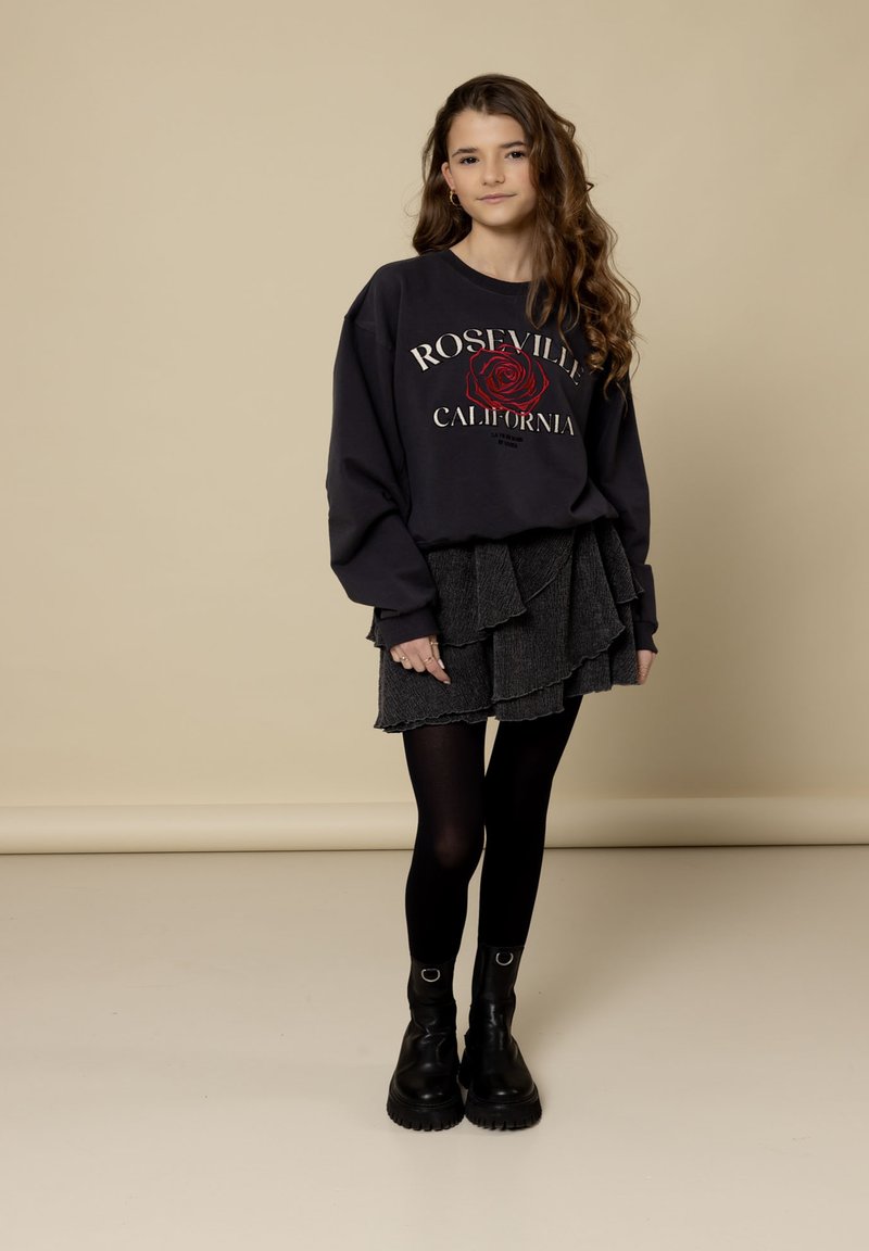 Tienermeisje met lang krullend haar, gekleed in een zwarte sweatshirt van Roseville, Californië, met een gelaagde zwarte rok, zwarte panty en zwarte laarzen tegen een beige achtergrond.