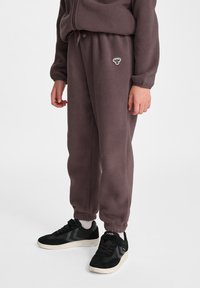 Fleece-Jogginghosen in dunklem Mauve mit elastischem Bund und Bündchen, versehen mit einem kleinen gestickten Logo auf der linken Seite. Kombiniert mit schwarzen Sneakern.