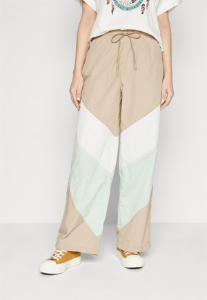 Pantalons de jogging beige et vert menthe avec un motif en chevron, fabriqués à partir d'un tissu léger, dotés d'une taille élastique et d'un cordon de serrage ; associés à des baskets beiges.