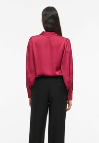 Kvinde med langt mørkt hår iført en langærmet rød satinbluse og sorte højtaljede bukser, vendt væk mod en hvid baggrund.