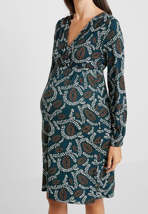 Femme enceinte portant une robe à manches longues, à motifs de feuilles et d'ovales bleus, marron et blancs sur un tissu vert foncé.