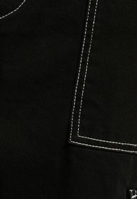 Detailní záběr černé tkaniny s bílým dvojitým stehem, který obrysuje roh kapsy na oděvu.