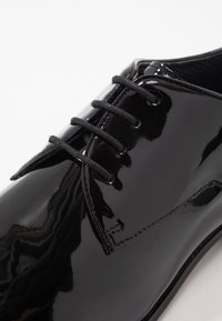 Scarpa in pelle verniciata nera con finitura lucida e liscia, dotata di cinque occhielli e lacci neri. Dettagli cuciti lungo i lati.