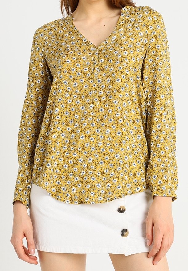 Femme portant un chemisier à manches longues fleuri jaune moutarde avec un col en V et une mini-jupe blanche à boutons.