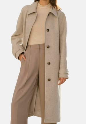 Femme portant un long manteau en laine beige boutonné, un pull à col roulé beige clair, et un pantalon taille haute à jambes larges couleur taupe.