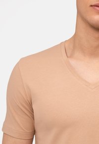 T-shirt beige con scollo a V realizzato in tessuto morbido e liscio. Maniche corte con vestibilità rilassata e fine cucitura lungo il collo.