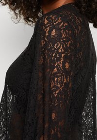 Černý krajkový top s květinovými vzory, průhlednou texturou a dlouhými rukávy, vyrobený z jemné látky a s otevřeným tkaním.