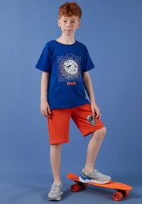 Garçon aux cheveux roux portant une chemise bleue à thème lunaire et un short orange, debout avec un pied sur un skateboard orange sur un fond bleu.