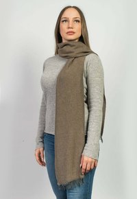 Brun texturerad halsduk med fransade kanter, hängande elegant, ihop med en ljusgrå tröja och blå jeans.