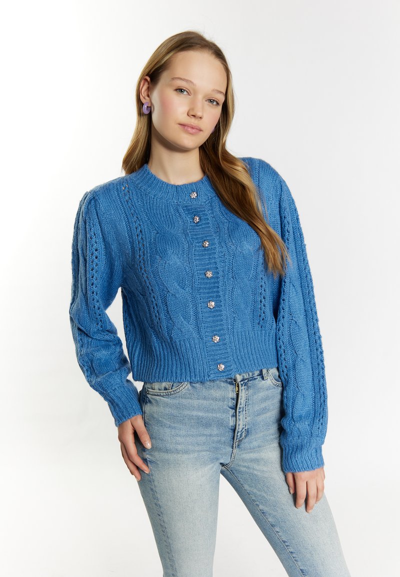 Jersey corto de punto azul con diseño de trenza, puños acanalados y botones similares a perlas en el frente. Combinado con jeans celestes.